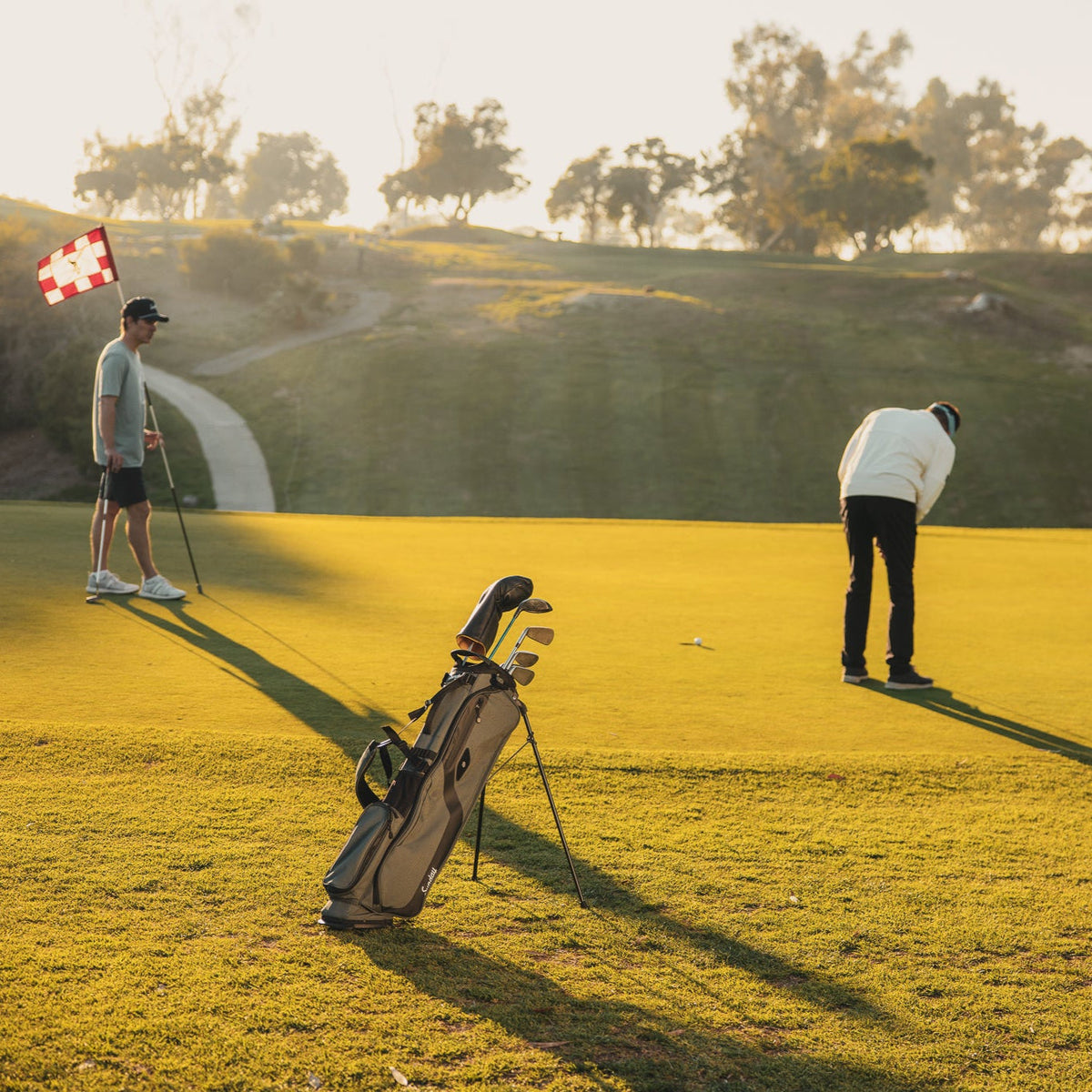 A standing El Camino Golf bag in midnight green on golf course with two golfers on the green putting 